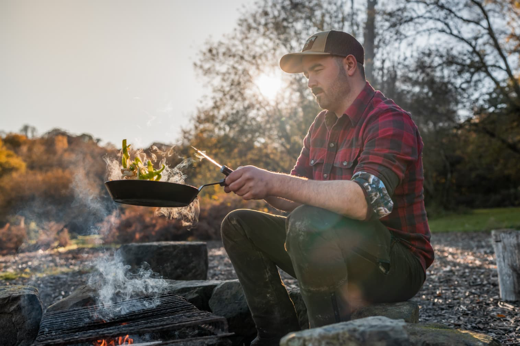 Cai Ap Bryn cooking over a campfire with a skillet at golden hour